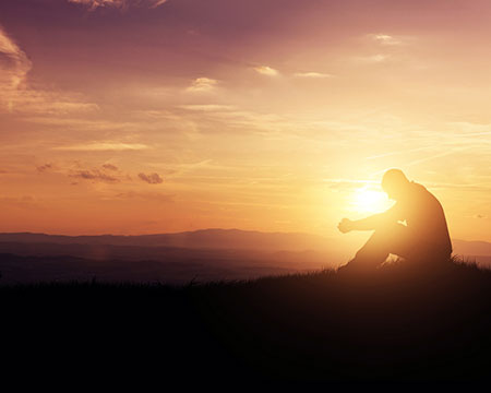 Catholic man bows his head in prayer with sunset in background